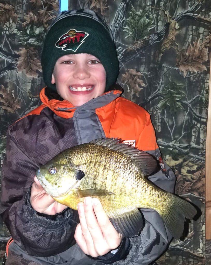 A smiling child in winter clothing and a beanie holds a large sunfish with both hands, proudly posing as if they've just reeled in a walleye. The background features a camouflage-patterned wall.