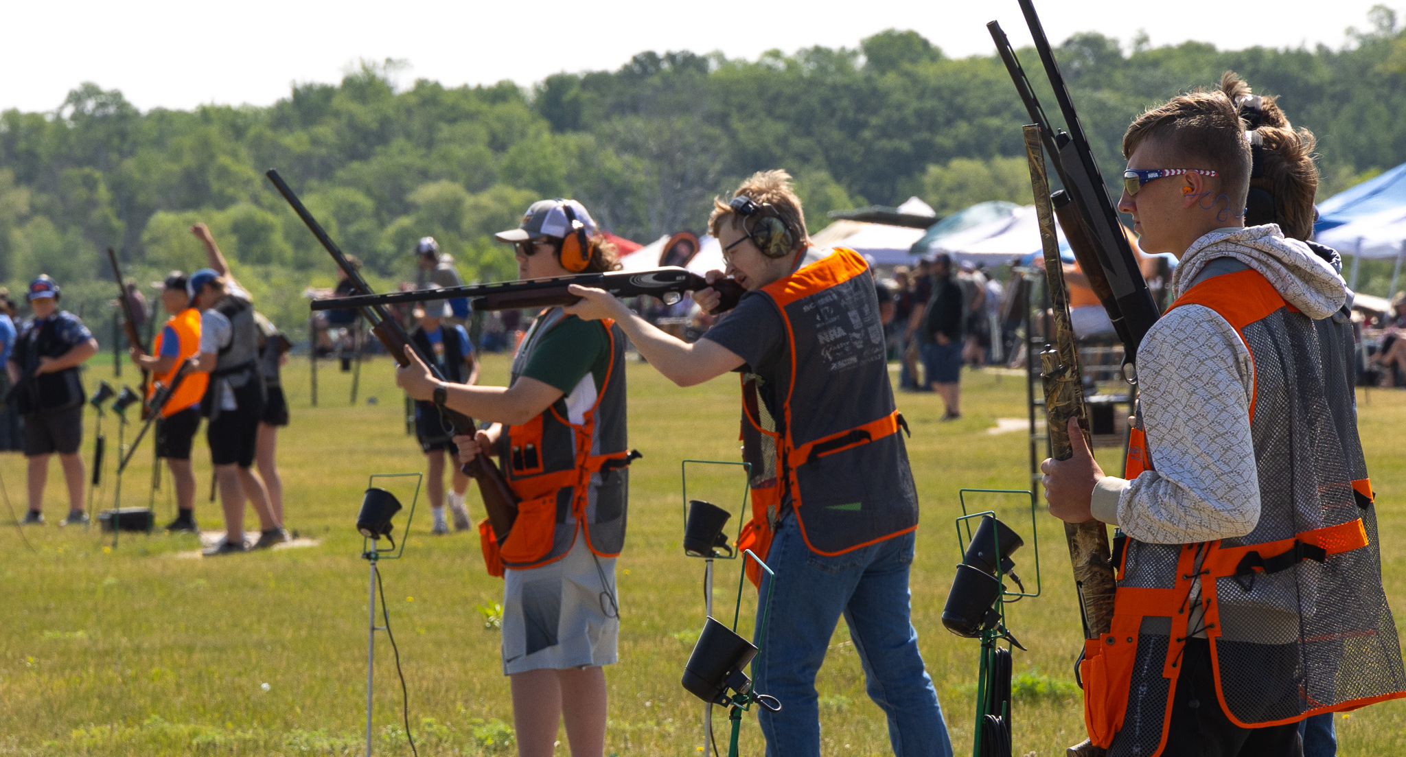 Several people wearing orange vests and ear protection stand outdoors aiming or holding shotguns at a shooting range, with others and equipment visible in the background on a sunny day.