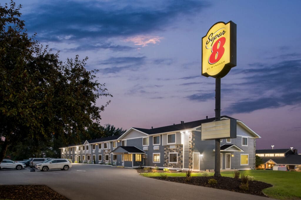 A Super 8 motel exterior at dusk features lit Super 8 signage, parked cars in front, and a tree framing the left side. The partly cloudy sky glows with subtle evening light.