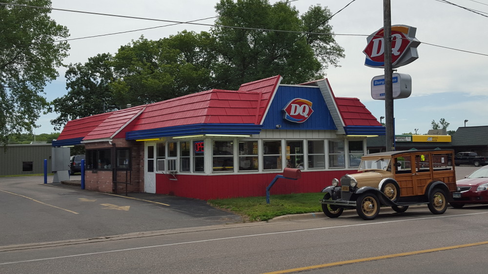 A Dairy Queen North restaurant with a red roof and blue trim sits near a road. An antique car is parked in front, and the DQ sign is visible above the building, with trees and other buildings in the background.