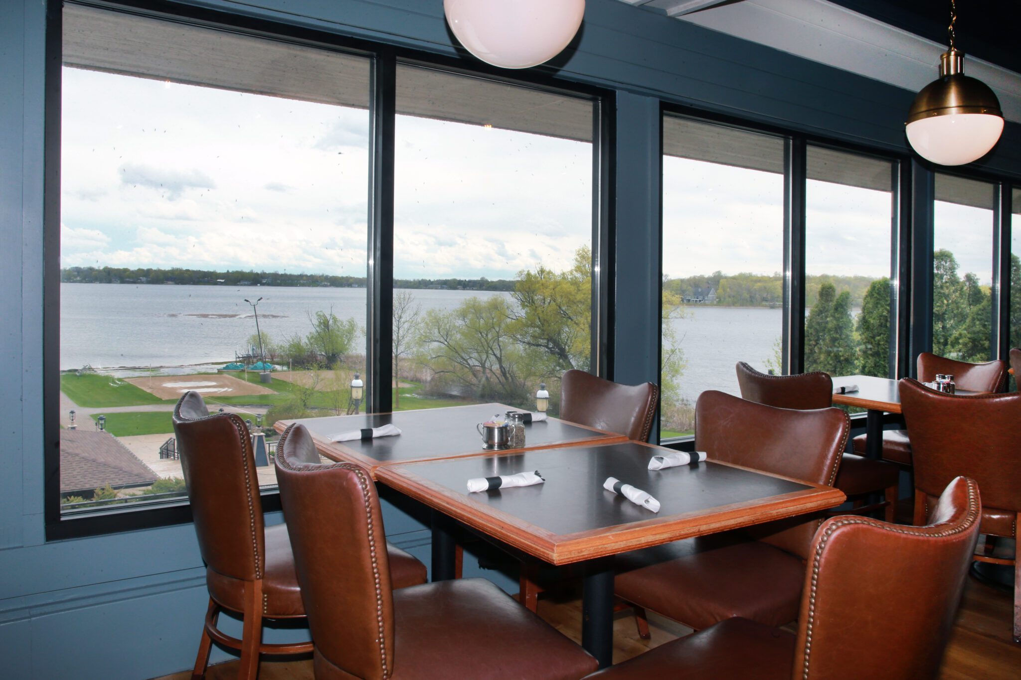 A restaurant dining area with brown leather chairs and set tables sits beside large windows at 2100 Chophouse, offering a scenic view of a lake, trees, and a cloudy sky outside.