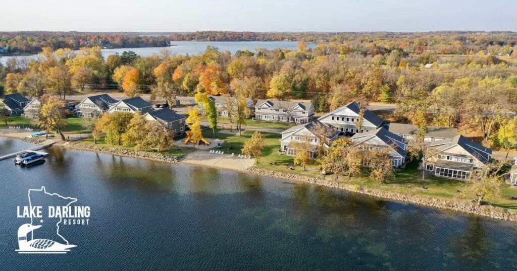 Aerial view of Lake Darling Resort reveals lakeside cabins nestled among autumn trees with vibrant foliage. The calm lake hugs the shoreline, with a dock in view. The "Lake Darling Resort" logo rests in the bottom left corner.