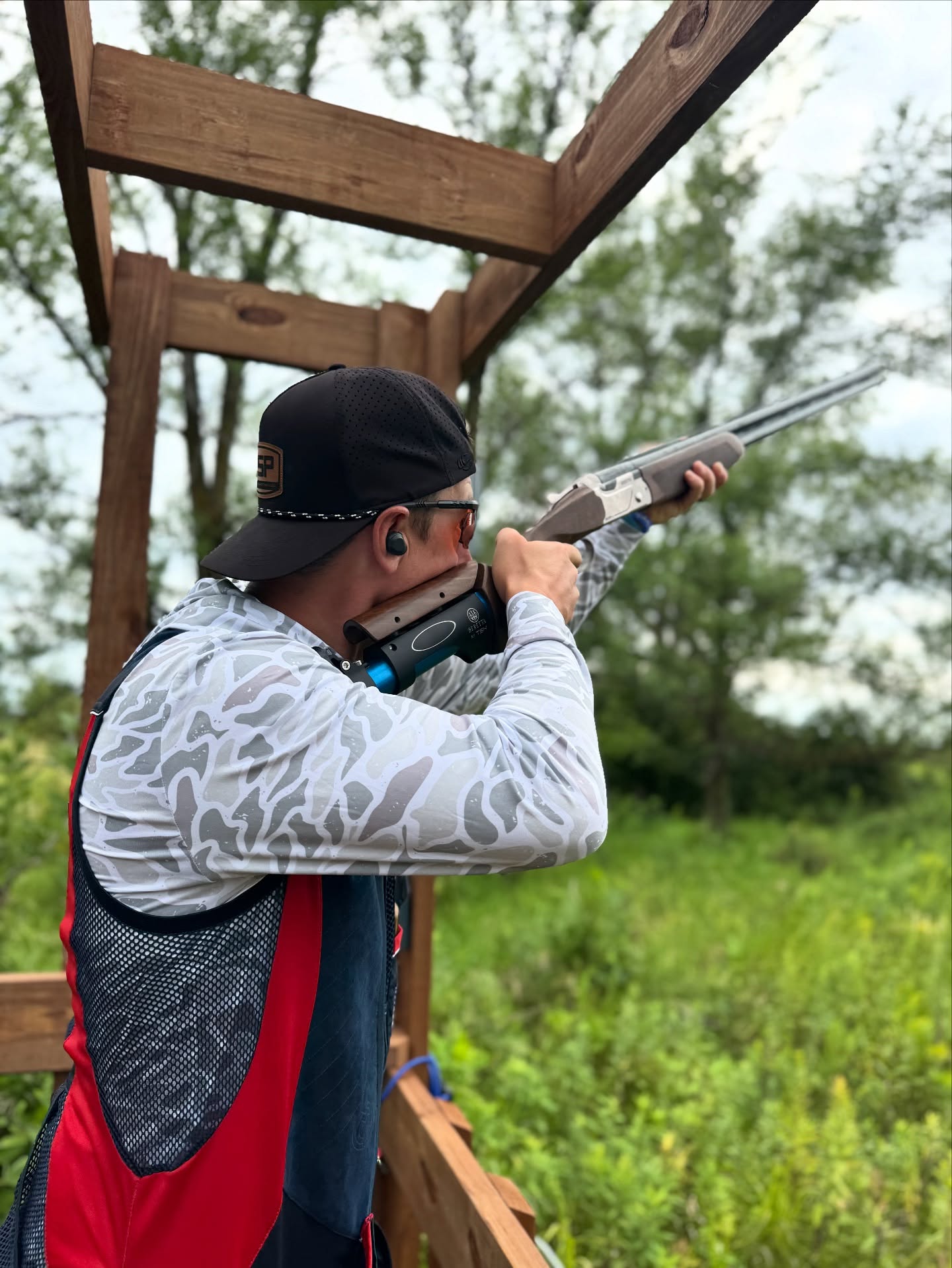 A person wearing a cap, sunglasses, and ear protection aims a shotgun outdoors at Alexandria Shooting Park, standing under a wooden structure surrounded by greenery and trees.
