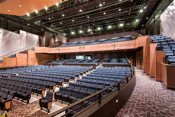 Spacious, modern auditorium at Alexandria Community Education Performing Arts with tiered seating, blue cushioned chairs, and contemporary lighting. Multiple seating levels and patterned carpet complement the large stage area at the front.