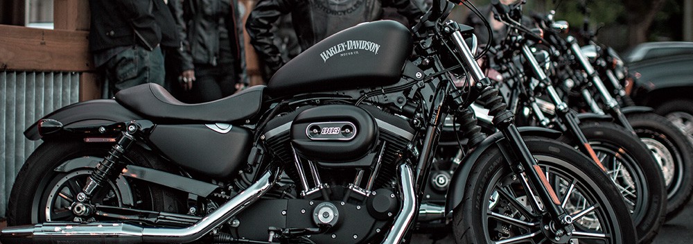 A row of black Apols Harley Davidson motorcycles parked outdoors, with several people nearby dressed in dark clothing. The focus is on the closest motorcycle, highlighting its details and logo clearly.