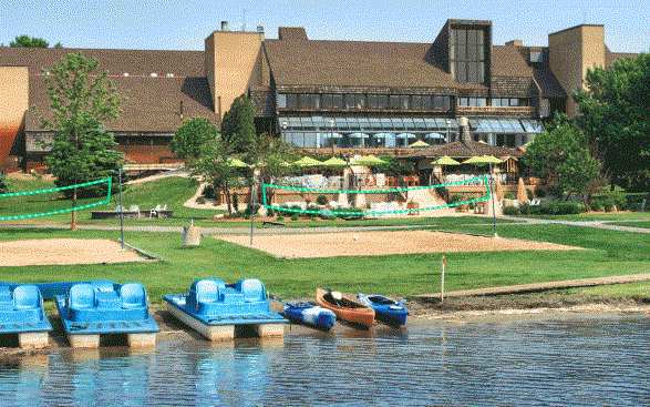 A lakeside resort with paddle boats and kayaks by the shore, sandy volleyball courts, green lawns, and a large lodge building with outdoor seating and umbrellas—enjoy all these amenities with Rentals at Arrowwood Resort.