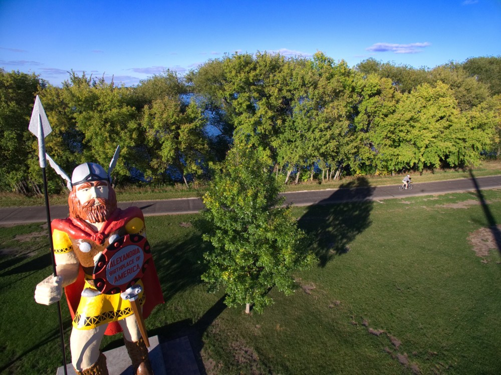 A large Viking statue with a spear and shield stands on grass near a row of trees and a paved path in Big Ole Park Alexandria MN; two people ride bicycles in the background under a clear blue sky.