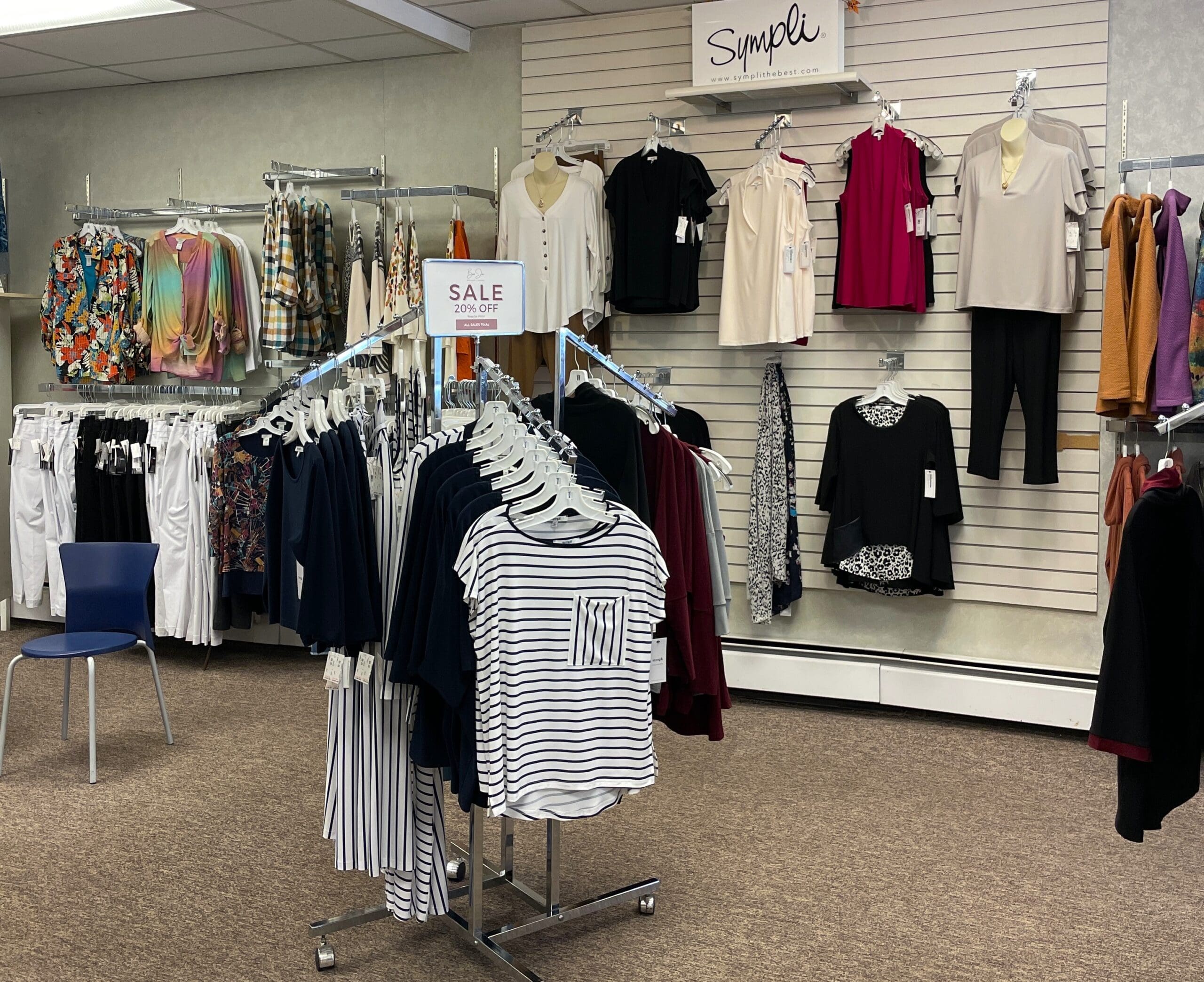 A clothing store display at Bon Jos Boutique features various women's tops and pants on racks and wall hooks. A "SALE 20% OFF" sign sits atop a rack of striped and solid shirts. The store has neutral walls and a blue chair.