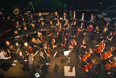 A large orchestra, like the Central Lakes Symphony Orchestra, sits on stage, viewed from above, with musicians playing various instruments. A conductor stands on a podium facing the ensemble—capturing the essence of live music in Alexandria MN.