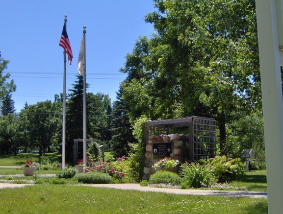 Two flagpoles with American and Illinois state flags stand in Curtis A Felt Memorial Park, a sunny green space with blooming flowers, trees, a stone sign surrounded by plants, and a trellis structure behind the sign.