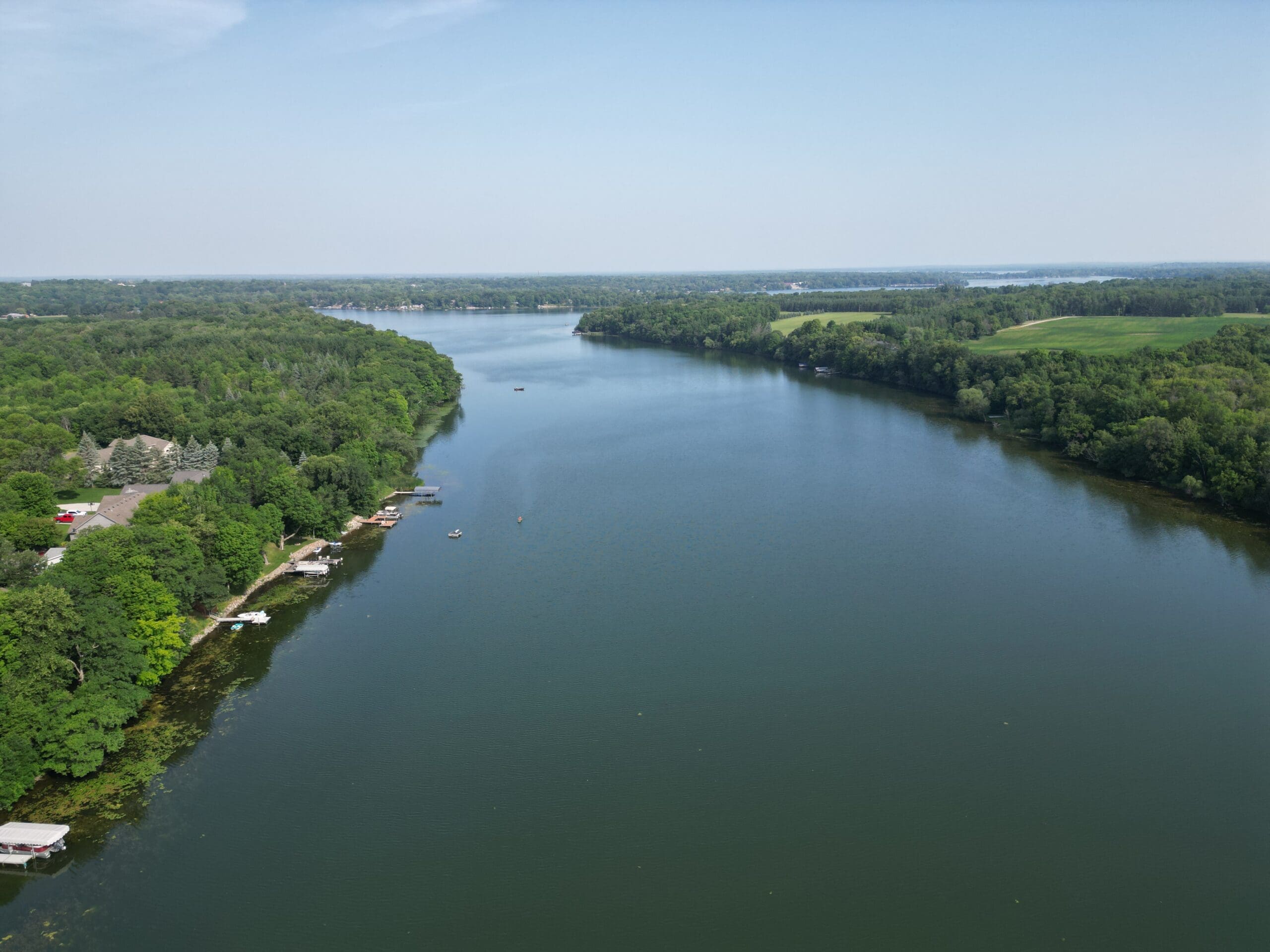 A wide river, reminiscent of Lake Victoria's serene beauty, curves gently through a lush, green landscape with trees lining both banks. Small docks and boats are visible along the left shore under a clear blue sky.