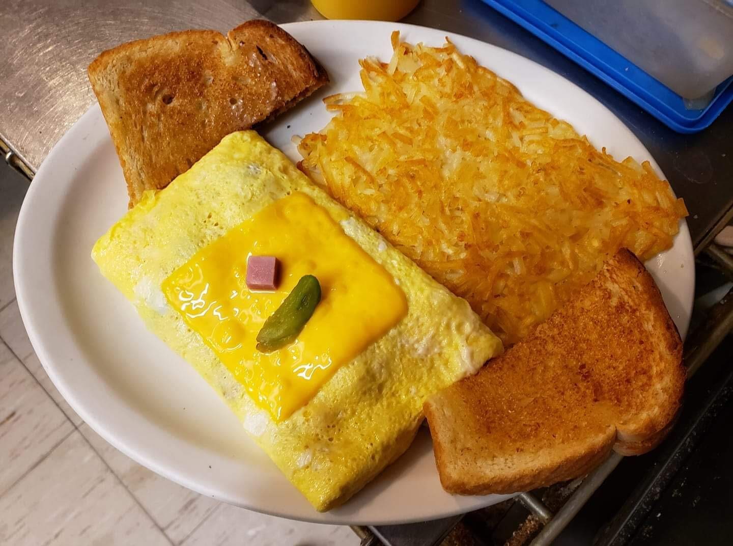 A Downtown Diner breakfast plate featuring two slices of toasted bread, a rectangular omelet topped with melted cheese, diced ham, and a jalapeño slice, served alongside golden hash browns.