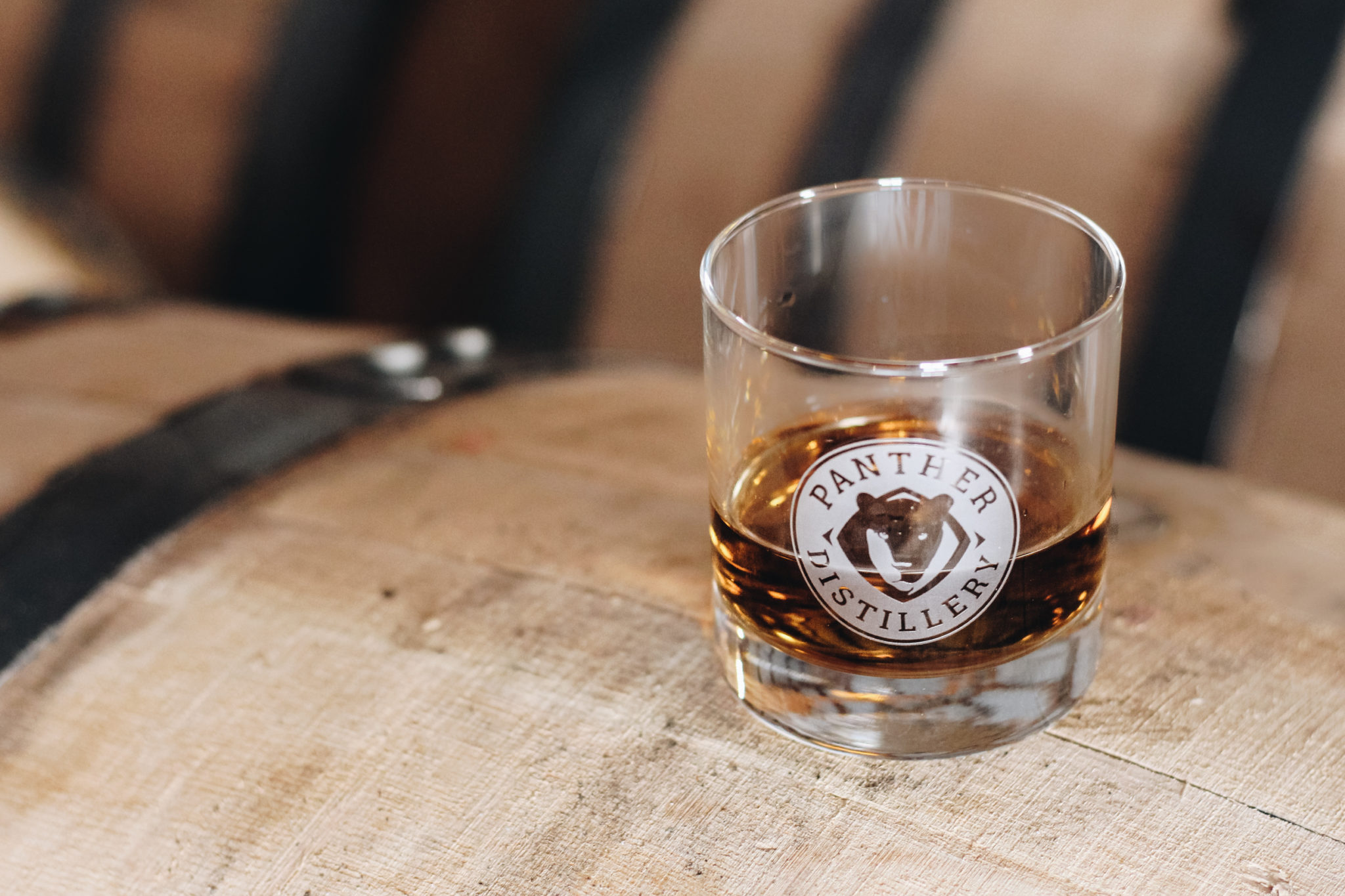 A glass with the Panther Distillery logo, filled with dark liquor, sits on top of a wooden barrel adorned with black metal bands in the background, capturing the authentic spirit of Panther Distillery.