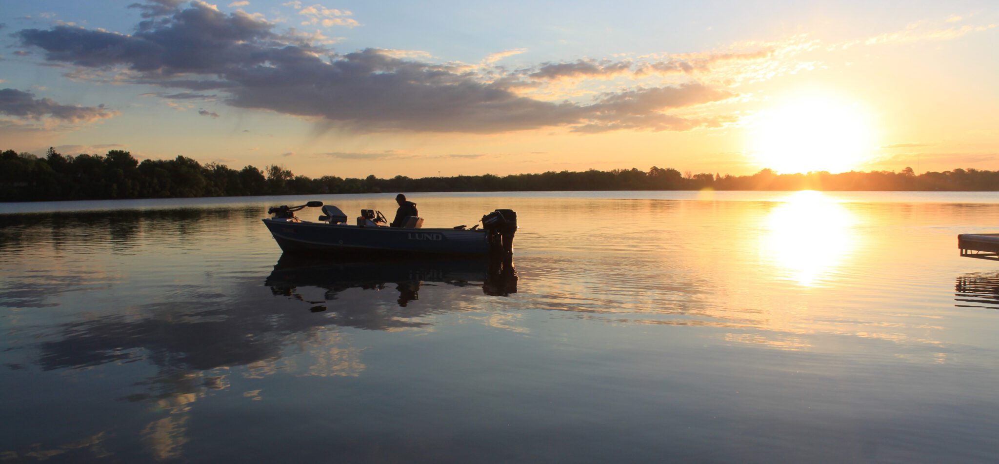 A small boat with two people floats on Lake Agnes at sunset, the sky glowing orange and reflections shimmering on the water. Trees line the horizon in the background.