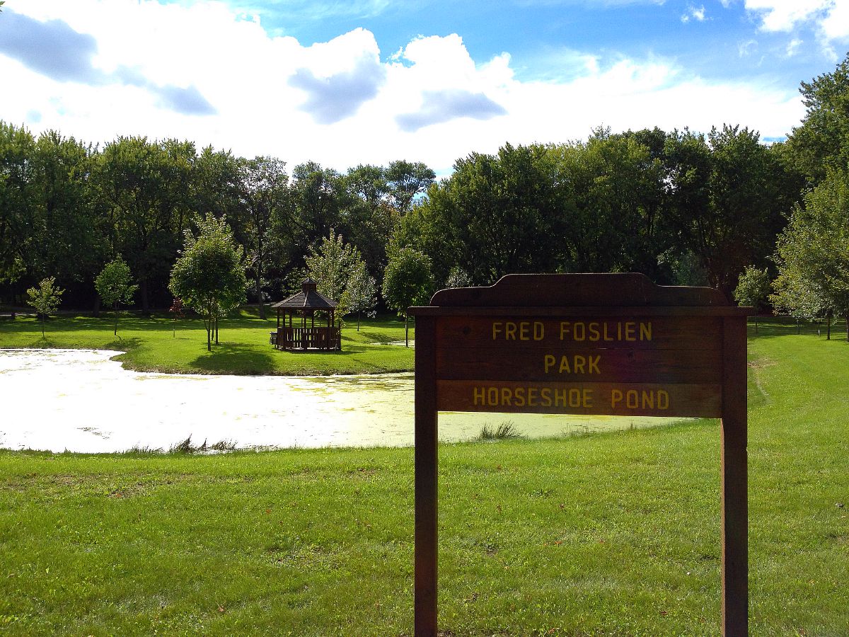 A wooden sign reading "Fred Foslien Park, Horseshoe Pond" stands on green grass by a small pond. In the background of Fred Foslien Park, trees and a gazebo are visible under a partly cloudy sky.