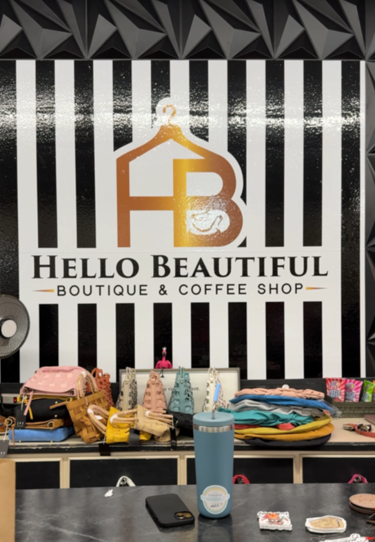 Hello Beautiful. A boutique counter displays bags, accessories, and a tumbler before a black-and-white striped wall with a gold “Hello Beautiful” logo and the text: “Hello Beautiful Boutique & Coffee Shop.”.