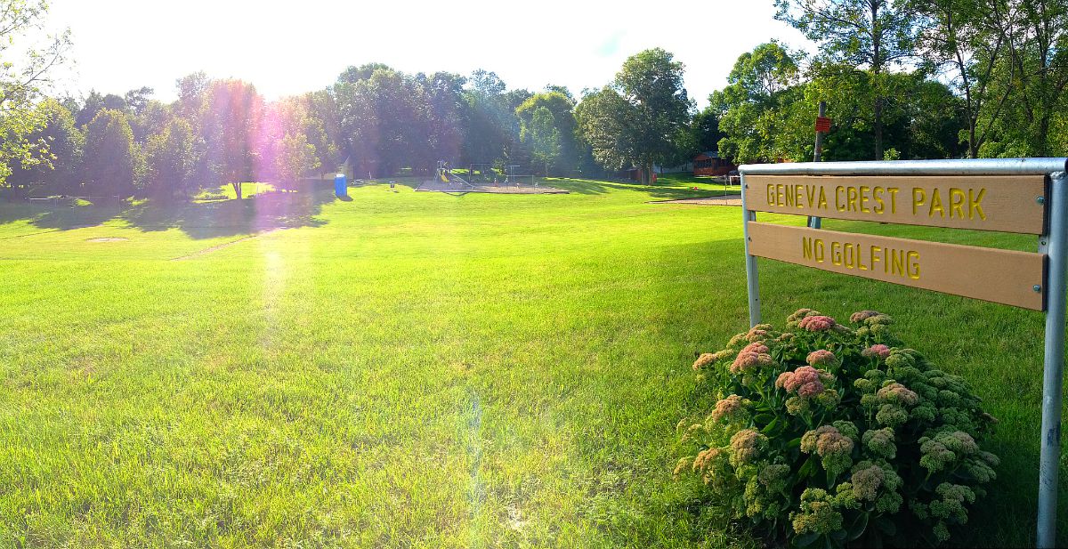 A sunlit park with lush green grass and trees. In the foreground, a wooden sign reads "GENEVA CREST PARK" and "NO GOLFING" beside flowering plants. Geneva Crest Park appears spacious and serene.
