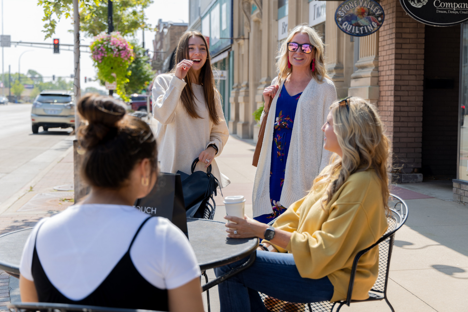 Four women enjoy a sunny day outside at a sidewalk café in downtown Alexandria; two sit at a table with coffee while two stand nearby, smiling and chatting. Cars and shops line the background street.