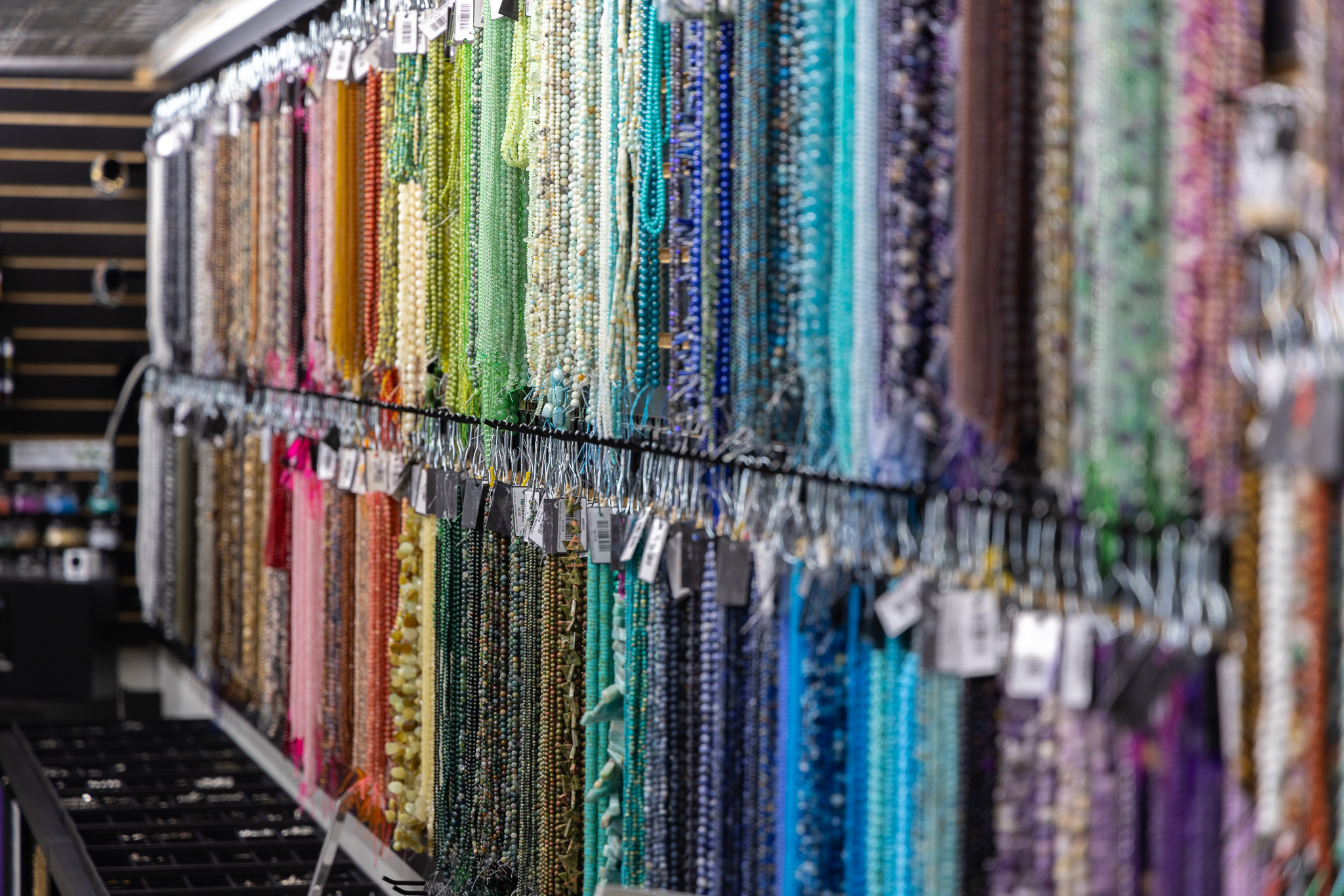 Strings of colorful beads in greens, blues, purples, pinks, and browns hang neatly organized on display racks at iTry Studios, creating a vibrant and inviting craft or bead store atmosphere.
