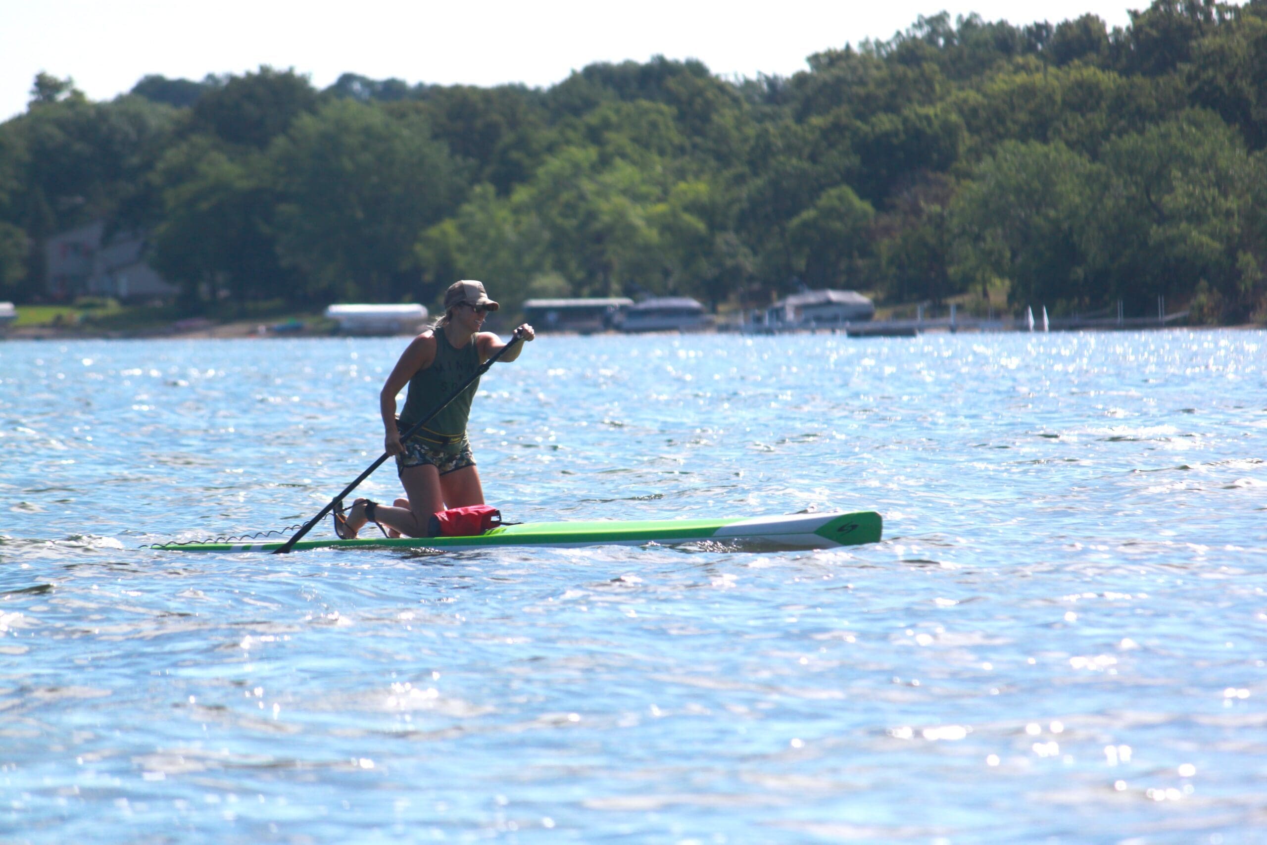 A person wearing a cap and sunglasses kneels on a paddleboard, paddling across sunlit Lake Ida with trees and boats visible in the background.