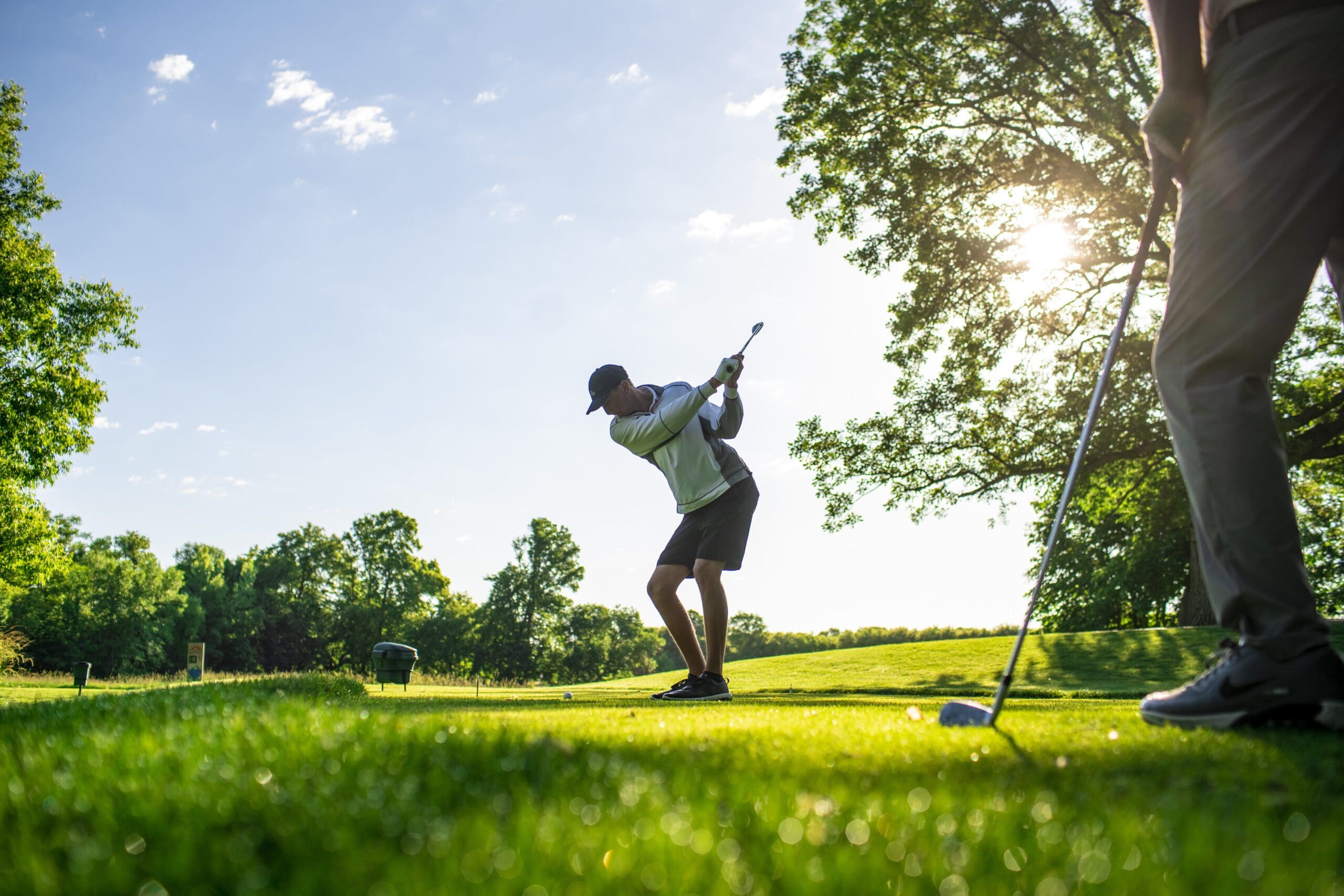 A golfer is mid-swing on the lush, green course at Geneva Golf Club under a clear sky, with sunlight streaming through the trees. Another person stands nearby, watching.