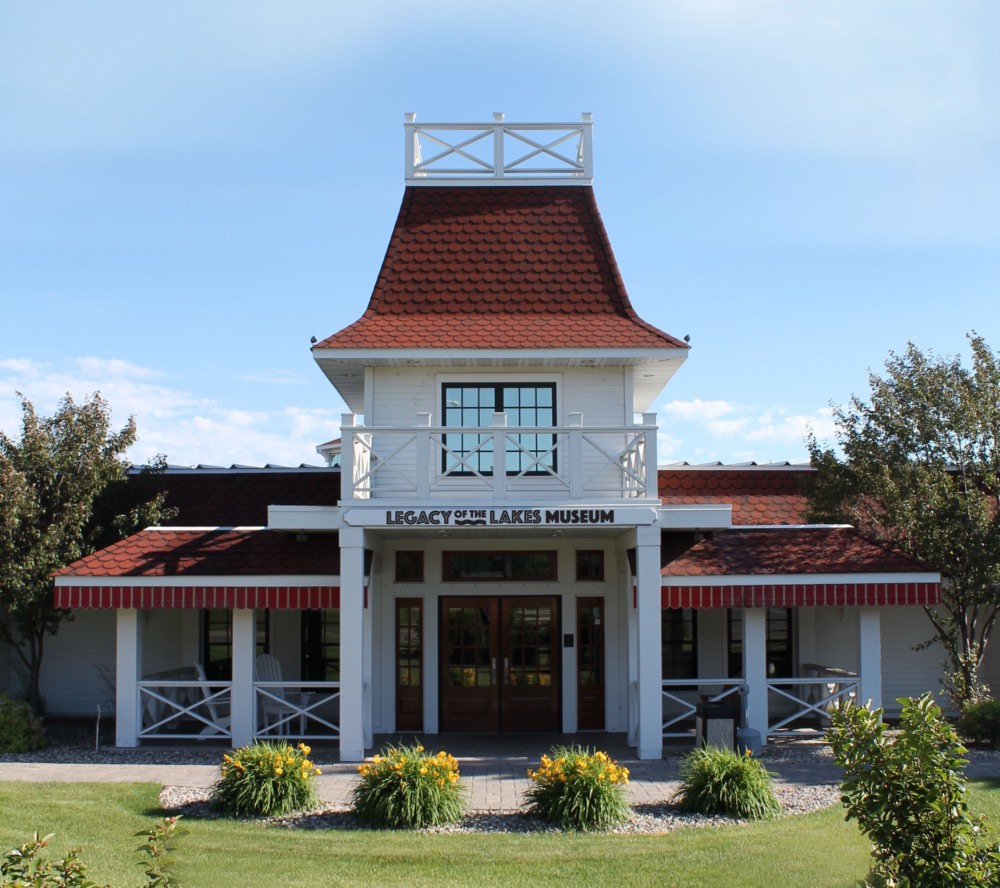 The Legacy of the Lakes Museum is housed in a white building with a red roof, featuring a central tower, front porch, and landscaping with yellow flowers and trees beneath a clear blue sky.