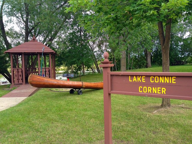 A brown wooden sign reads "Lake Connie Corner" near a gazebo and a large wooden canoe displayed on the grassy shores of Lake Connie, all surrounded by trees.