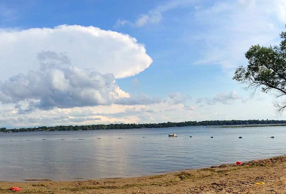 A sandy beach leads to the calm waters of Lake Darling, with a distant treeline on the horizon. The sky is partly cloudy, featuring a large billowy cloud. A few buoys and a floating object rest on the lake’s surface.