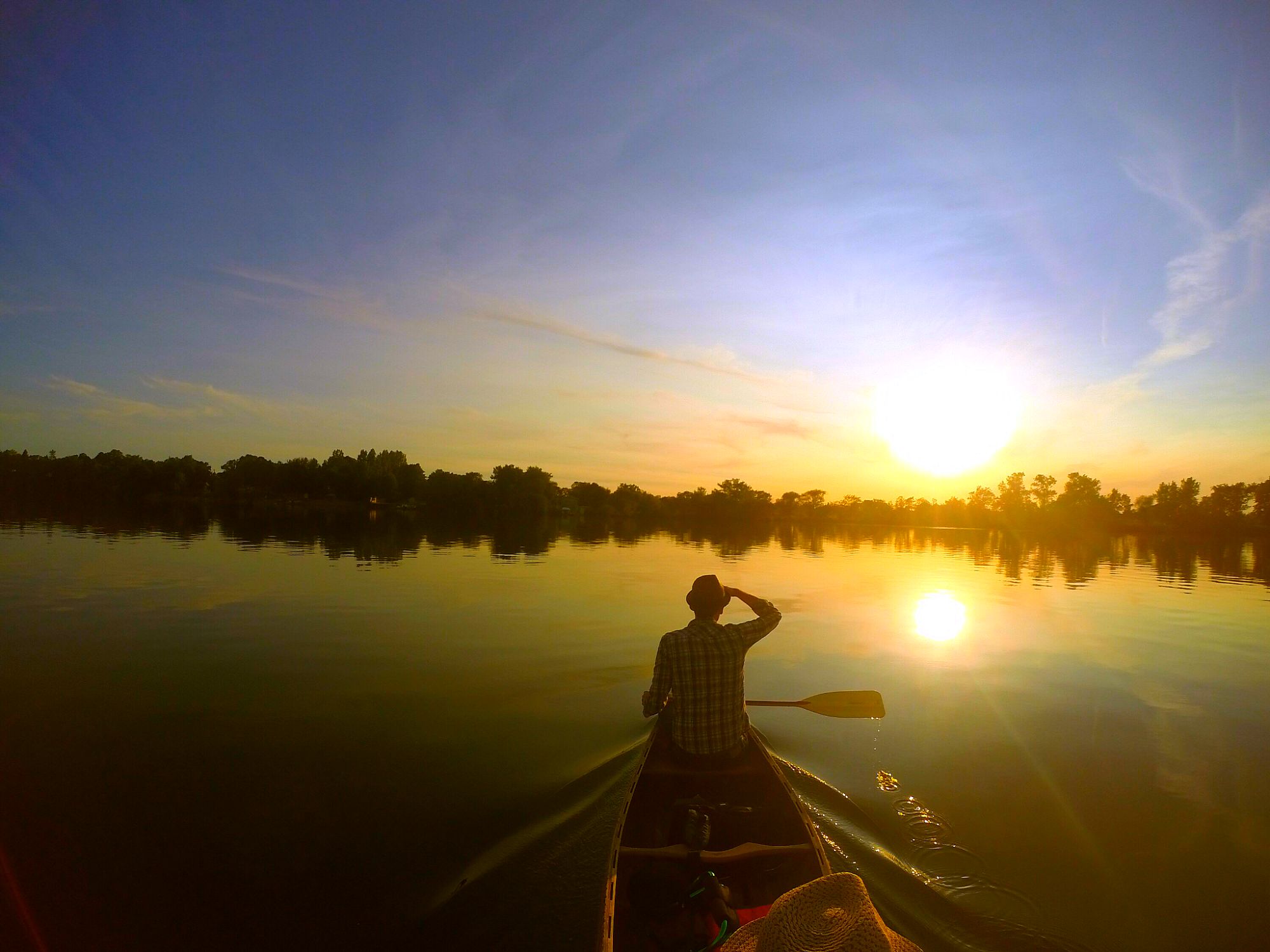 A person paddles a canoe on calm Lake Henry at sunset, with trees silhouetted along the shore and the sky glowing in shades of orange and blue.