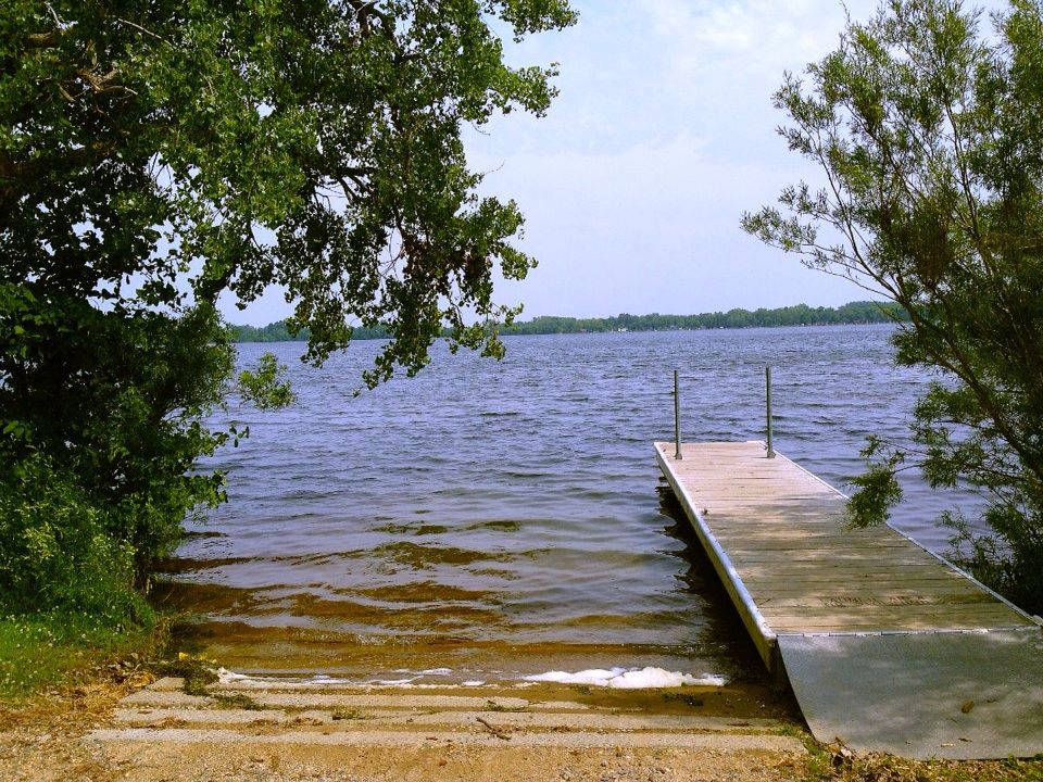 A small wooden dock extends into a calm lake reminiscent of Lake Louise, surrounded by trees and greenery under a partly cloudy sky. Steps lead down to the water’s edge, bordered by lush foliage on both sides.