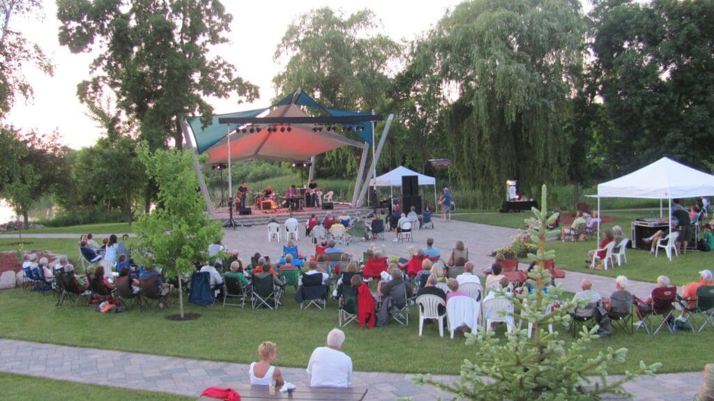 Outdoor concert with a band performing on a stage under a canopy, surrounded by trees. An audience sits on chairs and blankets on the grass, enjoying the music. White tents are set up nearby on a sunny day.