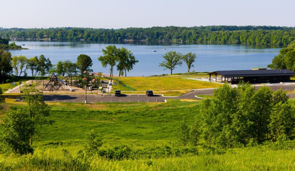 Lake Brophy County Park features a playground, picnic shelter, and parking lot with a few cars, all surrounded by grass and trees. The park overlooks a calm lake with a forested shoreline in the background.