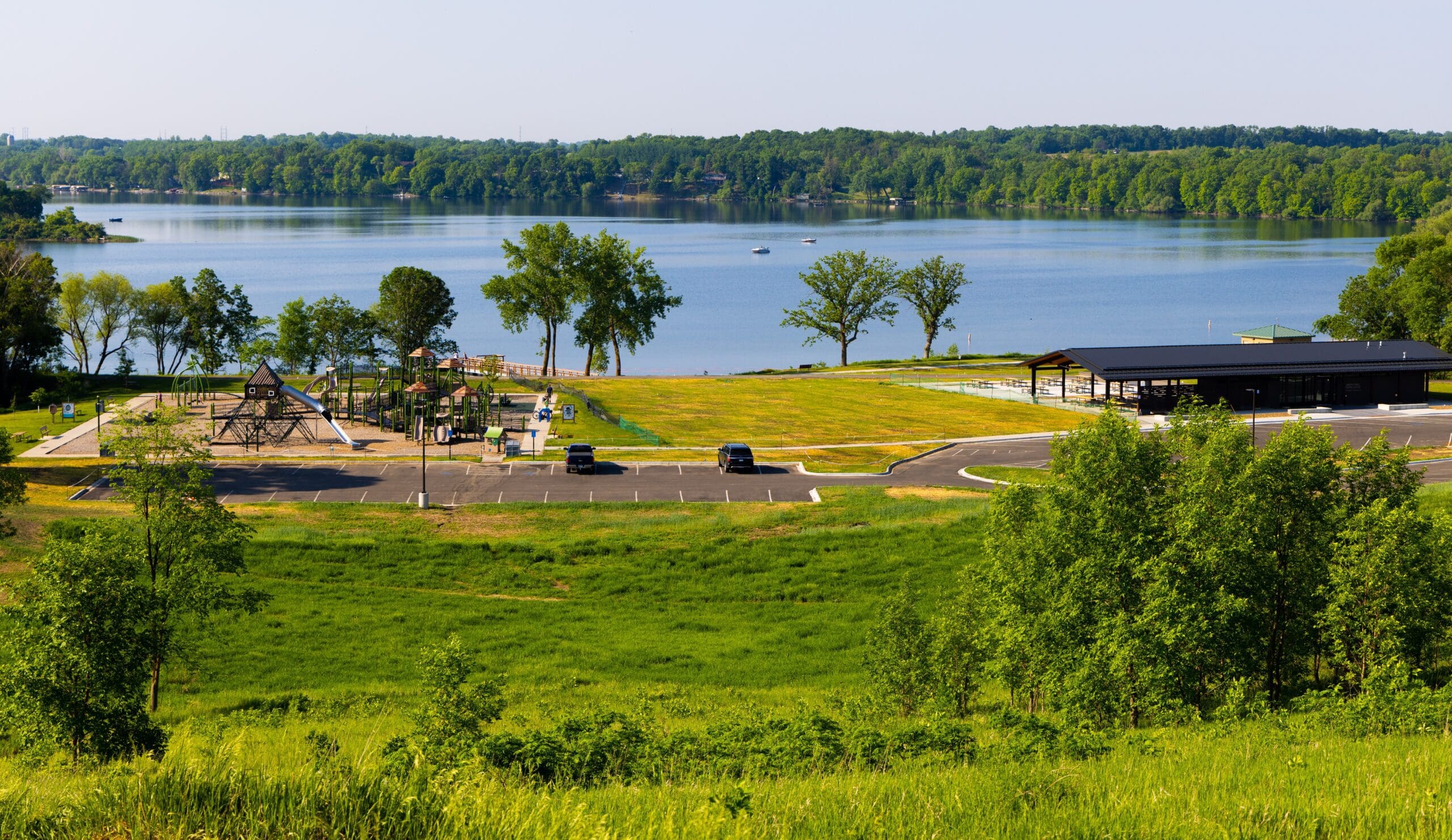 Lake Brophy County Park features a playground, picnic shelter, and parking lot with a few cars, all surrounded by grass and trees. The park overlooks a calm lake with a forested shoreline in the background.