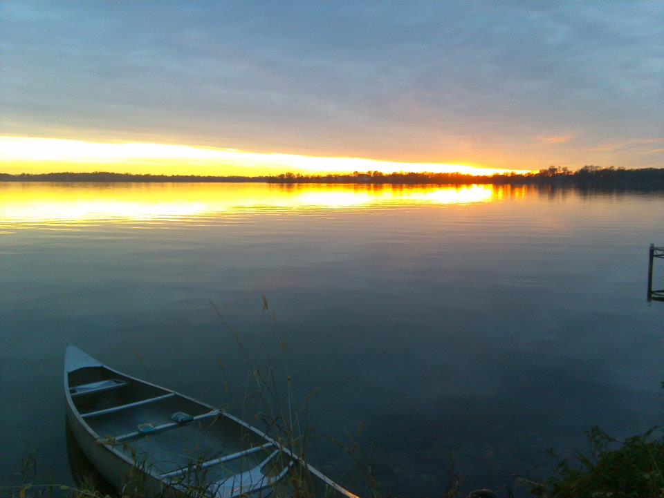 A canoe rests on the shore of Maple Lake at sunset, with golden sunlight reflecting off the calm water and a clouded sky overhead.