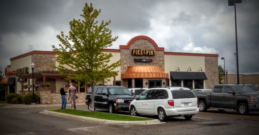 A bar and grill restaurant named "Pike & Pint" features a stone and stucco exterior, several parked cars in front, and three people standing near the entrance on a cloudy day.