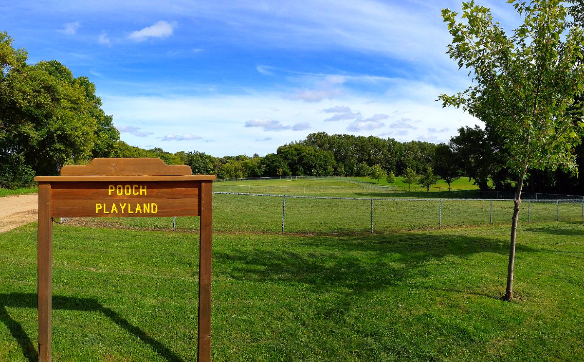 A wooden sign reading "Pooch Playland Dog Park" stands at the entrance of a large, grassy, fenced-in dog park surrounded by trees under a blue sky with scattered clouds.
