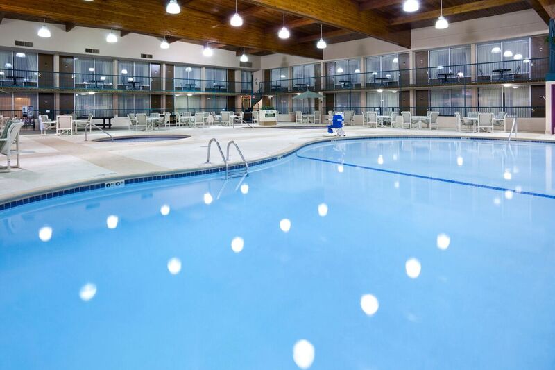 Large indoor swimming pool with clear blue water at Holiday Inn, surrounded by lounge chairs and tables. Overhead lights reflect on the water, while glass-walled hotel rooms line the two-story building around the pool.