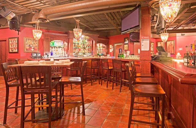 A cozy bar, Willy T's, with high wooden chairs and tables, warm lighting from hanging lamps, shelves of liquor bottles behind the bar, red walls, and framed artwork creates an inviting atmosphere.