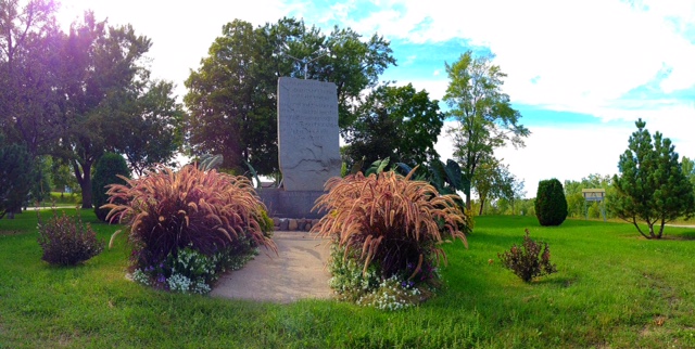 A stone monument stands in Runestone City Park, surrounded by ornamental grasses, small flowering plants, and trees beneath a partly cloudy sky.