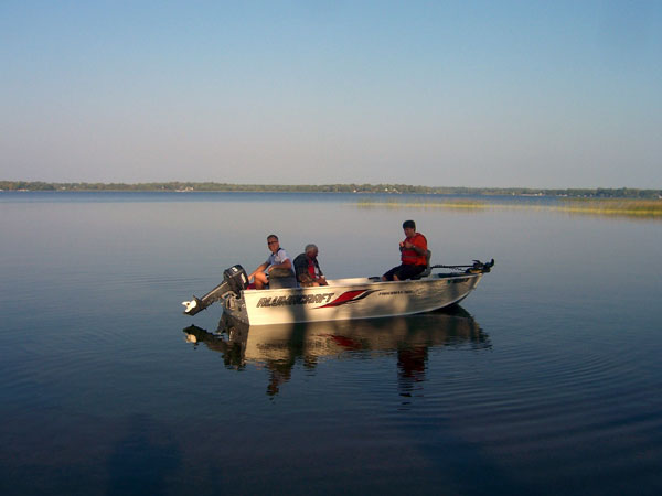 Three people sit in a small motorboat labeled "WARRIOR BOAT" on calm, reflective water under a clear blue sky, enjoying a scenic view of the distant shoreline and trees—perfect for those visiting the nearby RV campground.