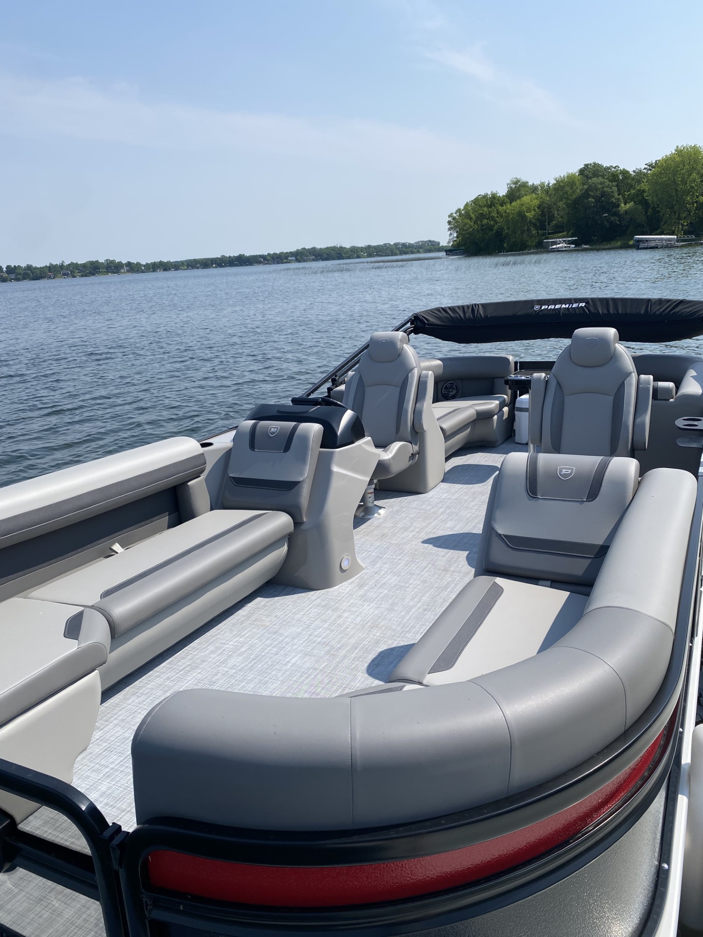 A spacious pontoon boat with gray and beige cushioned seating floats on a calm lake near Betsy Ross Resort, surrounded by trees and a distant shoreline under a clear sky.