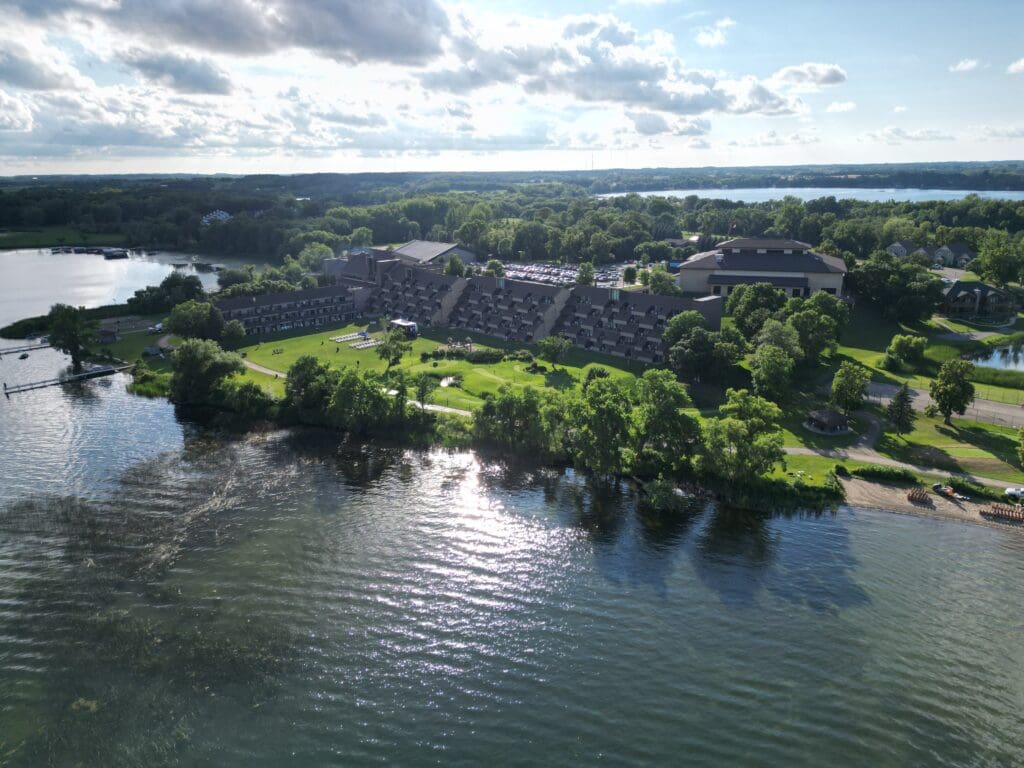 Aerial view of Arrowwood Resort, a large building complex surrounded by trees, located next to a lake with calm water. The sky is partly cloudy, and sunlight reflects off the water’s surface.
