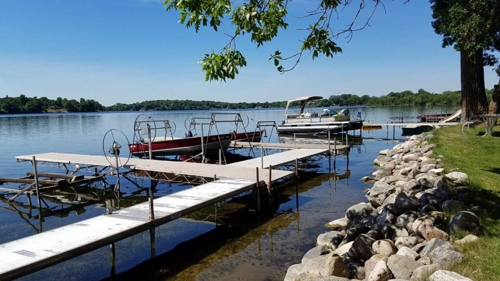 A peaceful lakeside scene at Vacationer's Inn, with docks extending over the water, two boats moored, and a rocky shoreline bordered by green grass under a clear blue sky. Trees are visible in the distance across the lake.