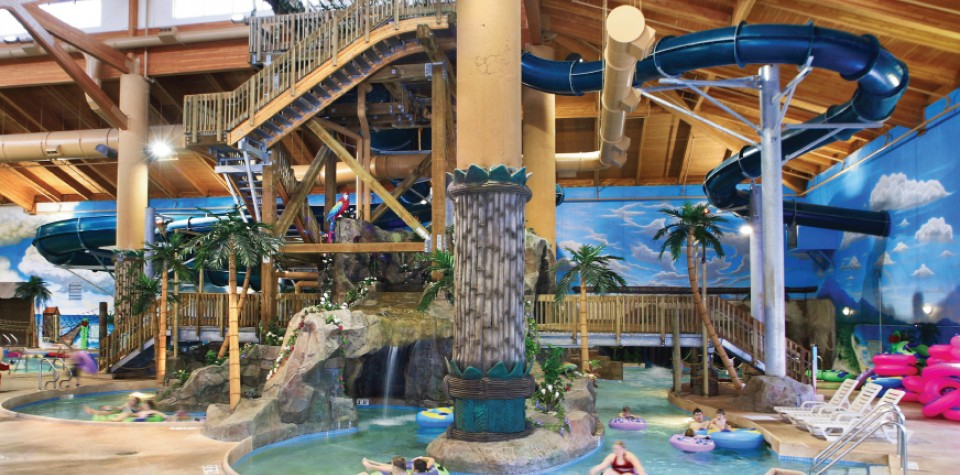 Big Splash Indoor Waterpark features a large blue waterslide, wooden walkways, fake palm trees, and people floating in inner tubes. Bright, tropical-themed decor and a high wooden ceiling complete the fun atmosphere.