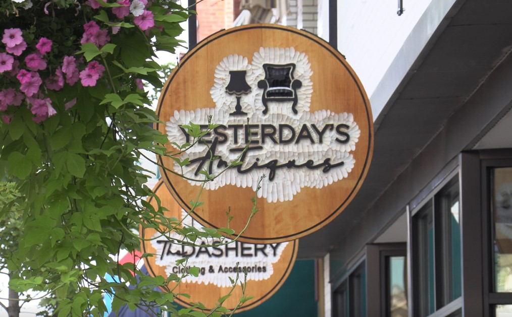 A wooden shop sign reads "Yesterday's Antiques" with images of a lamp and armchair above the text. Partially visible below is another sign for "Yesterday's Haberdashery Clothing & Accessories." Flowers and greenery hang nearby.