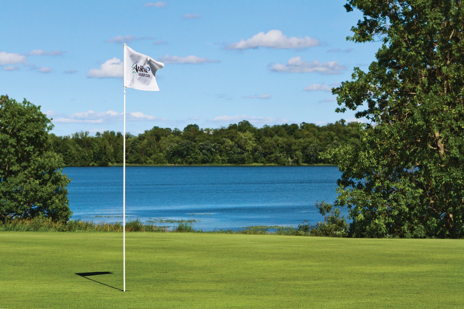 A white golf flag on a green course at Atikwa Golf Course stands near a calm blue lake, surrounded by trees under a partly cloudy sky.
