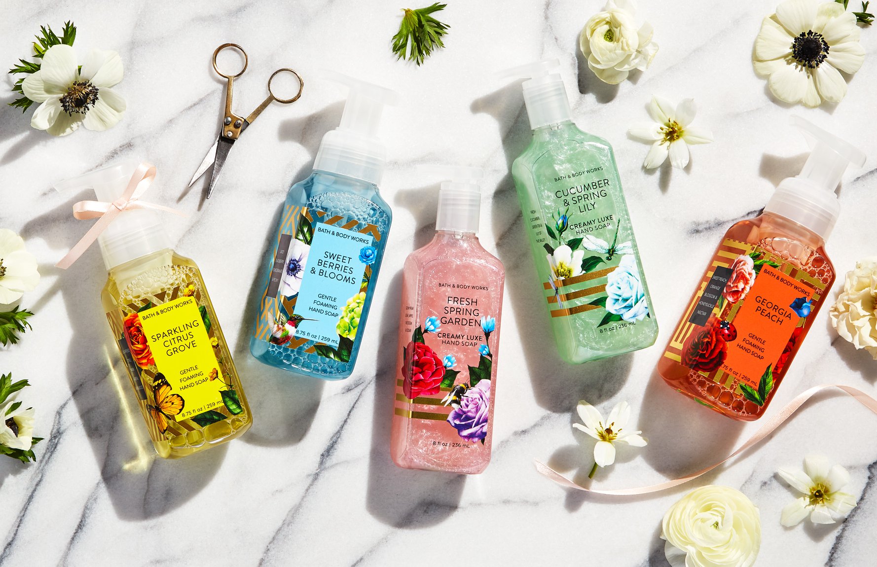 Five colorful hand soaps with floral labels, including Bath & Body Works, are arranged on a white marble surface, surrounded by white flowers, gold scissors, and a pink ribbon. Scents featured include peach and cucumber.