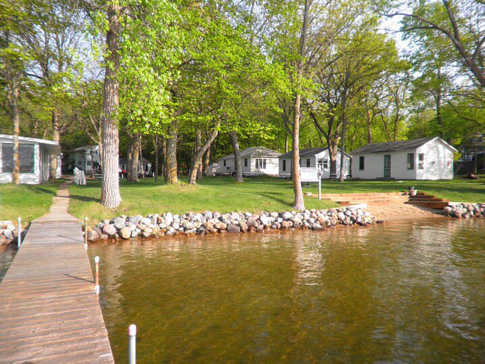 A wooden dock extends into a clear lake at Berg's Resort, with a sandy shore. Several white cottages are nestled among tall, leafy trees, and green grass and rocks line the shoreline on a sunny day.