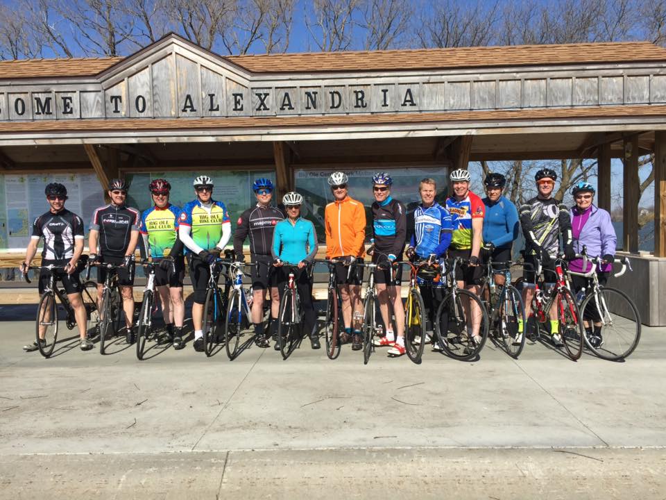 A group of 13 cyclists from the Big Ole Bike Club, wearing helmets and cycling gear, stand with their bicycles in front of a wooden shelter that says "Welcome to Alexandria" on a sunny day.