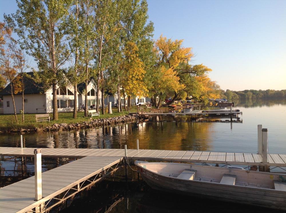 A calm lake with wooden docks and a rowboat in the foreground at Brophy Lake Resort, lined with trees showing green and yellow leaves. Houses and benches sit along the waterfront under a clear sky.